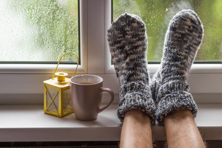 Mans feet wearing knitted wool socks near the hot cup of tea with yellow lantern on windowsill at cozy autumn home.の写真素材