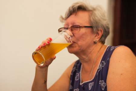 Smiling caucasian senior woman with gray hair holding and drinking glass of light beer in craft czech brewery.の写真素材