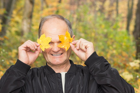 Happy senior man Holding and covering her eyes with two yellow maple leaves in the autumn city park. Active seniors.の写真素材