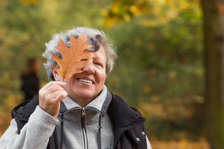 Happy senior woman with toothy smile covering her eye with brown oak leaf in the autumn city park....