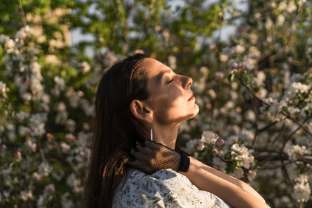 Attractive young woman with closed eyes enjoying fresh air and sun rays in blooming apple tree garden. She holding brown hair with arms and relaxing in the spring park. beauty in nature concept.の写真素材
