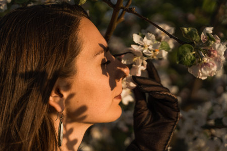 Young brunette woman enjoys by blooming apple trees and sniffs it flowers with closed eyes in spring garden. She holds tree branch with white flowers and smells it, close up. Seasonal allergy concept.の写真素材