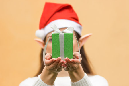Young woman in santa hat, with elf ears holding green gift box with bow in front of her. Creative...
