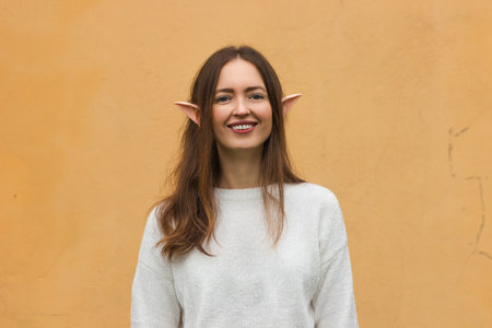 Portrait of happy young woman with toothy smile and elf ears in white sweater isolated on yellow background. Christmas or New Year party look. Preparation for winter holidays masquerade event.の写真素材