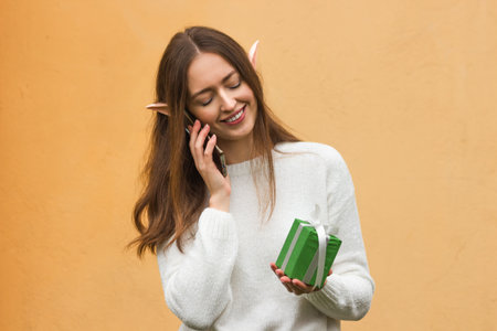 Smiling young woman with elf ears talking on mobile phone and holding green gift box in hand,...