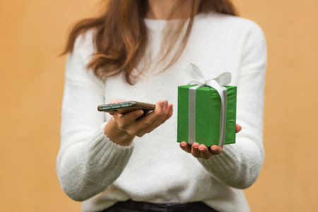 Woman's hands holds smartphone and green gift box with bow, isolated on yellow background. Female in white sweater using mobile app and shopping online. New Year and Christmas holidays concept.の写真素材