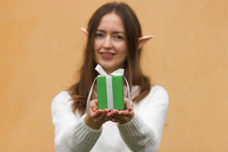 Young woman in white sweater with elf ears holding green gift box with bow in front of her. yellow...