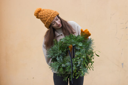Young woman in knitted ginger hat and mittens looking at the Christmas wreath. Florist holding handmade rustic wreath with pine and fir branches, mistletoe berries on light background. xmas home decorの写真素材