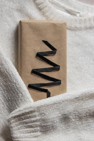White warm sweater with sleeves hugs a gift box wrapped in kraft recycled paper, decorated with black ribbon in shape of Christmas tree. Plastic free, eco Xmas wrapping idea.の写真素材