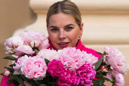 Portrait of caucasian blonde woman in crimson dress with bouquet of pink peonies. Happy young female holding bunch of flowers. St. Valentine's Day, International Women's Day holiday concept.の写真素材