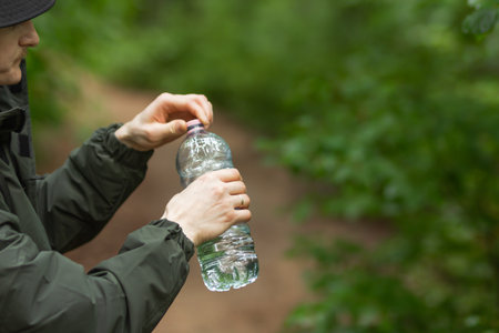 Young caucasian man opening plastic cap of bottle to drink water, close-up. Tired man resting after hiking in spring forest and holding bottle of water in hands. Plastic problem and ecology concept.の写真素材