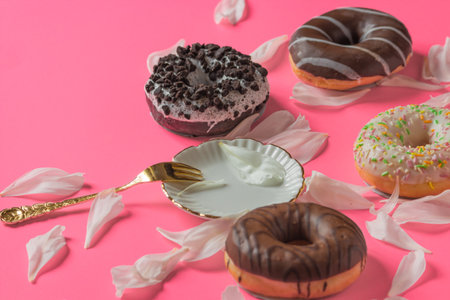 Different donuts coated with a chocolate and white frosting isolated on a pink background with white petals of peony flower, vintage saucer and golden fork. National Donut Day concept. springtime.の写真素材