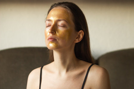 Portrait of caucasian young woman using golden facial mask for moisturizing or peeling. Relaxed female with closed eyes applying golden skin cosmetic. Daily skin face treatment at home concept.の写真素材