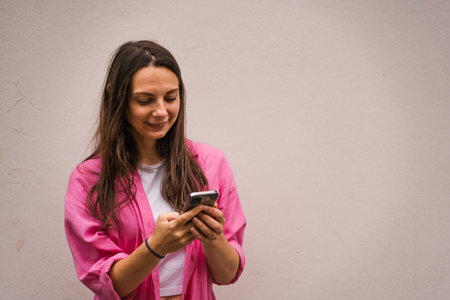 Portrait of a caucasian brunette smiling woman in casual pink shirt using smartphone. People and...