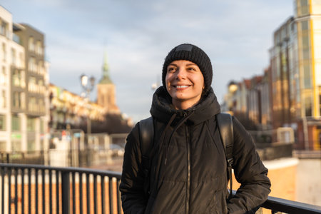 Smiling young woman in black jacket and warm hat traveling with backpack. Happy female walking on the city streetの写真素材
