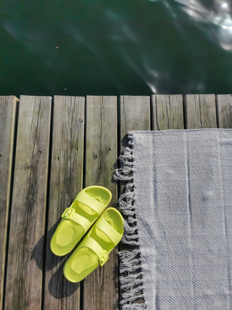 Womens pool sandals or flip flops in green neon color with beach rug on wooden floor, terrace beside deep lake water.の写真素材