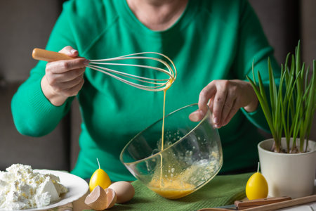 Elderly woman beats eggs and cane sugar with whisk in glass bowl. Prepare for making cheese pancakes or curd Easter.の写真素材