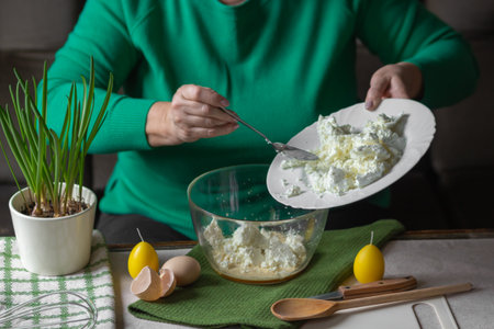 Senior woman adds spoonful of cottage cheese to the dough in glass bowl. Ingredients for cheese pancakes or curd Easter.の写真素材