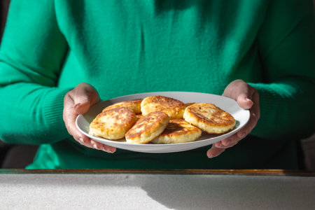 Old woman holds plate of tasty roasted cottage cheese pancakes - syrniki, ready to eat. Cooking healthy food concept.の写真素材