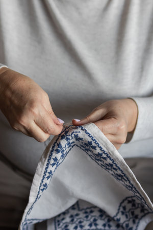 Senior womans hands embroidering a cross-stitch floral pattern on linen fabric with needle and blue...