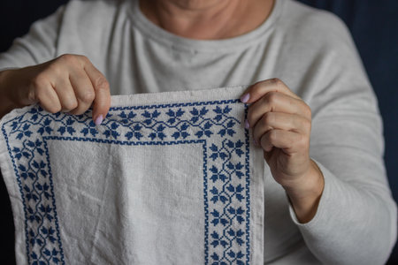 Senior womans hands showing an embroidery a cross-stitch floral pattern on linen fabric with blue...
