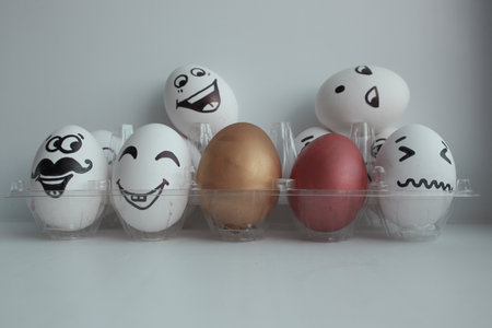 Eggs Easter white group in a transparent box with cheerful and mischievous faces having fun and standing on the heads of neighbors and two colored eggs in the middle.の写真素材