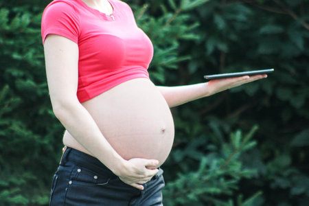Maternity concept. Girl in red on a nature background. Holds a tablet. In the profile. Photo for...