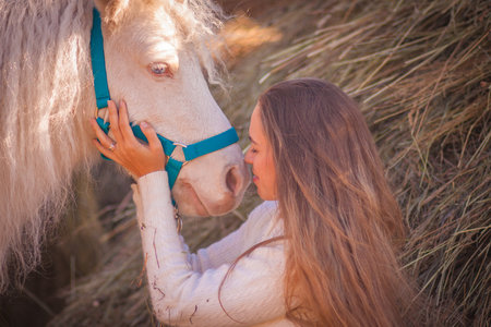 beautiful girl with white mane pony. resting in a haystack in the fall in the afternoon. horizontal orientation of the sheet. A cheerful girl with a smile talking with a pony nose to nose. communicatesの写真素材