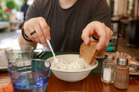 Close-up shows male hands holding spoon and dark bread above bowl of tzatziki. Natural light, traditional setting, salt and pepper shakers in frame.の写真素材