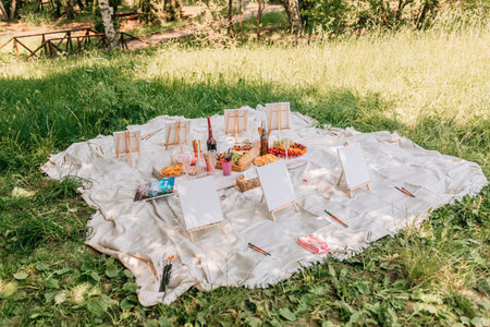 Large white blanket on grass featuring several easels with canvases, brushes, wine bottles, and vibrant fruits and snacks for painting gatheringの写真素材