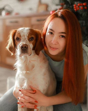 Portrait of young woman with straight copper hair embracing affectionate spaniel at cozy home interior. Natural daylight, gentle expression, holiday mood, wooden kitchen backgroundの写真素材