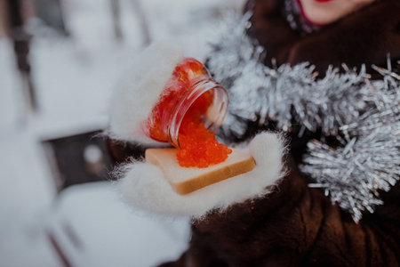 Close-up of female hands in soft mittens pouring red caviar onto bread slice. Snow background and fur coat create luxurious and festive winter atmosphere.の写真素材