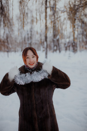 Cheerful woman in fur coat and patterned scarf standing outdoors in snow, holding shiny tinsel decoration. Festive winter atmosphere, natural daylight.の写真素材