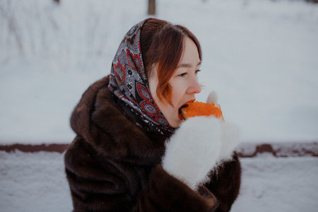 Close-up of female model in traditional headscarf and fur coat enjoying sandwich with bright orange...