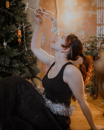 Playful female celebrating New Year in stylish black dress, creative humor with tinsel used as festive drink. Warm light and Christmas decor create cozy atmosphere.の写真素材