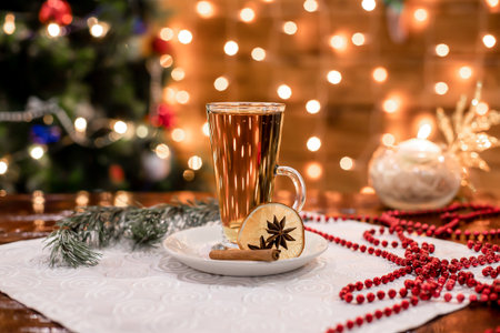 Hot herbal drink in tall glass surrounded by festive decor and blurred lights. Cinnamon stick and dried citrus create holiday aroma. Ideal image for winter drink promotions, menus,の写真素材