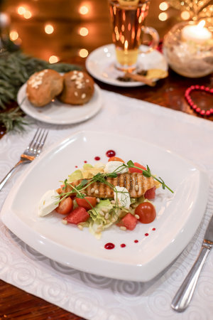 Close-up of grilled fish fillet salad with greens, mozzarella, cherry tomatoes, pine nuts, and sauce drops, served under warm festive lights.の写真素材