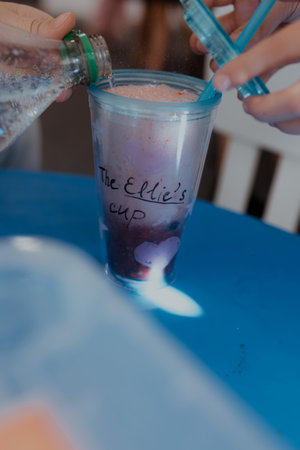 Sparkling liquid poured inside clear tumbler filled with berry mix creating fizzy drink reaction captured during kids craft drink activity with hands holding straws above blue table surface.の写真素材