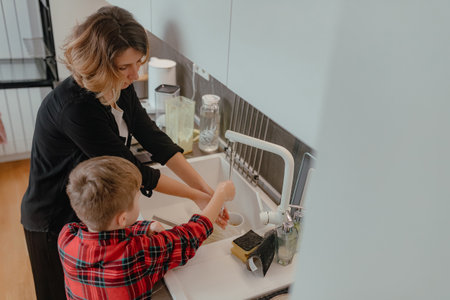 Adult woman helping young boy wash hands under running water inside bright home kitchen following cooking process with visible sink plus counter items around space.の写真素材