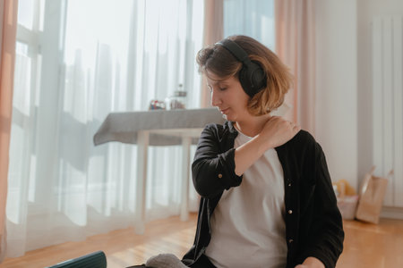 Woman massages shoulder while seated on blue exercise mat wearing wireless headphones inside bright home wellness space with warm daylight, relaxed expression, gentle stretch gesture, calm self-care moment.の写真素材
