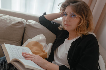 Bright living room scene showing woman wearing glasses reading book, soft window light, relaxed...