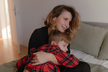Mother hugging child wearing red plaid pajamas during warm daylight inside cozy bedroom, relaxed posture, nurturing closeness, soft bedding, peaceful family moment captured naturally.の写真素材