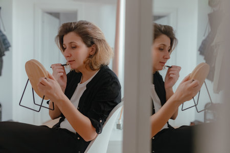 Woman applying lip liner while holding wooden mirror, seated near bright interior, soft natural...