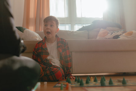 Boy expressing surprise during board game moment with wide open mouth, red plaid outfit, strong sun...