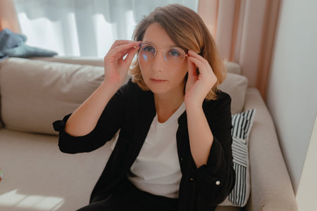 Woman adjusting eyeglasses while seated on beige sofa, soft daylight illuminating face, relaxed...
