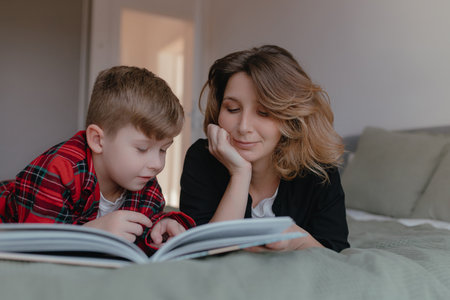 Soft light illuminates boy reading picture book beside mother, calm relaxed learning moment inside cozy bedroom environment, close interaction focused childhood education and gentle family bonding.の写真素材