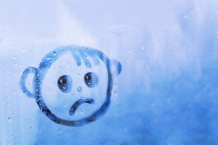 Foggy glass on blue window with drawn sad face concept photoの写真素材