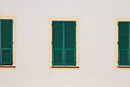 three green wooden window with shutters in yellow frame on white wallの写真素材