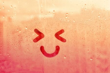Happy smile face with eyes narrowed painted on window flooded with raindrops on blur glass red yellow background in cityの写真素材