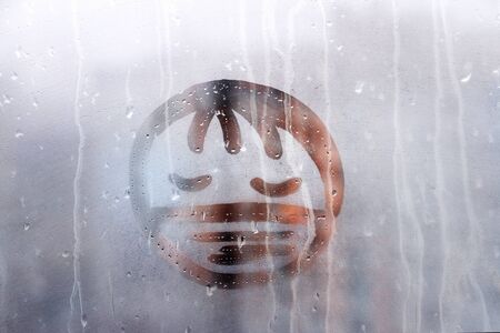 sad face with closed eyes in protective medical mask is painted in warm orange on a wet blue window...